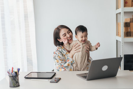 A little boy Asian baby in mother's arms, excited and joyful use computer laptop with video calls with father or long-distance relatives. Tech-Fueled Family Love Concept.の写真素材