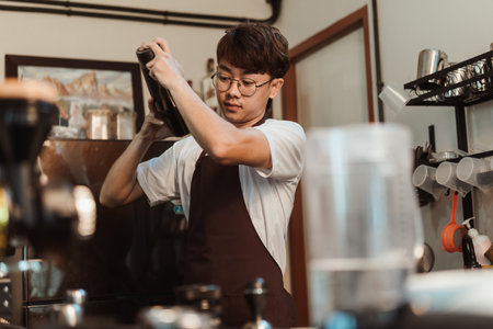Barista Asian man holds in hands steel shaker at coffee shop. Asian man barista in white t-shirt and apron working at coffee shop. SME business coffee shop concept.の写真素材