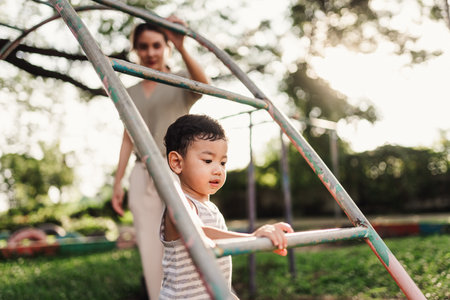 An Asian boy is playing on monkey bars in a playground while his mother stands nearby to help and ensure his safety. Active play and the joy of developing skills.の写真素材