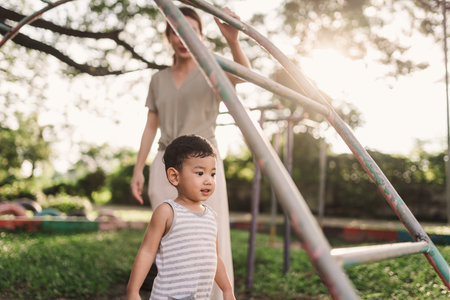 An Asian boy is having fun playing in a playground while his mother stands nearby to help and ensure...