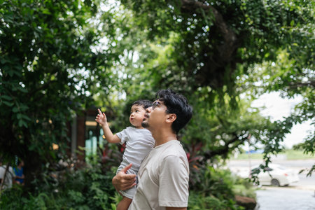 Asian father holding his son close as they look at something interesting in their backyard. The scene captures a peaceful moment of connection, and the joy of spending time together.の写真素材