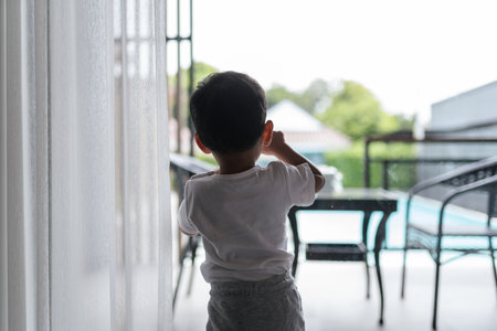 A little boy stands at a window, looking out of a window during quarantine. capturing a kid longing for the outside world. Pandemic and quarantine concept.の写真素材