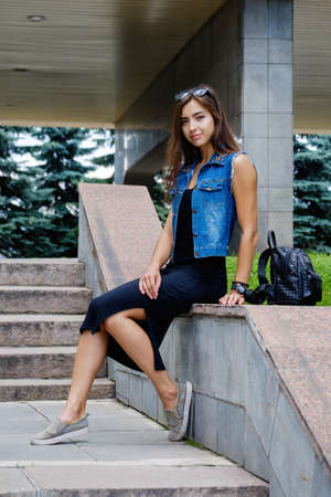 Beautiful sexy young brunette woman with long wavy hair thin slim figure perfect body and beautiful face make-up, wearing a black suit and vintage denim jacket. summer accessory, black backpackの写真素材