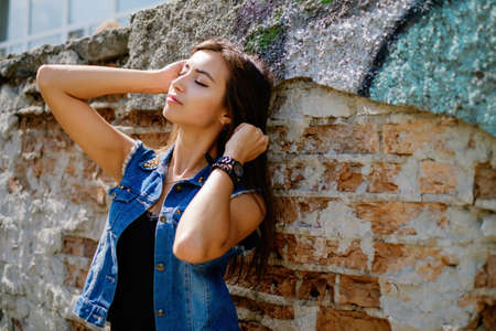 Beautiful sexy young brunette woman with long wavy hair thin slim figure perfect body and beautiful face make-up, wearing a black suit and vintage denim jacket. summer accessory, black backpackの写真素材