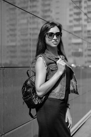 Beautiful sexy young brunette woman with long wavy hair thin slim figure perfect body and beautiful face make-up, wearing a black suit and vintage denim jacket. summer accessory, black backpackの写真素材