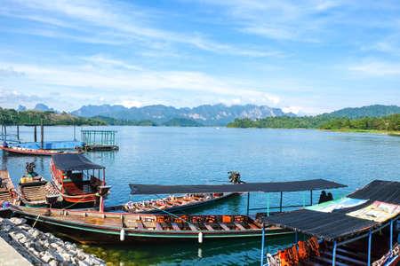 Wooden Thai traditional long-tail boat on a lake with mountains and rain forest in the background during a sunny day at Ratchaprapha Dam at Khao Sok National Park, Surat Thani Province, Thailandのeditorial素材