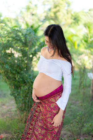 Young Asian beautiful pregnant woman standing in white dress touching her bellyの写真素材