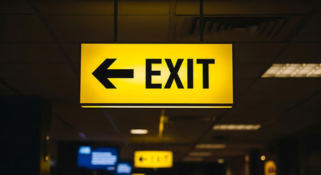 A brightly lit yellow exit sign with a black arrow pointing left hangs from a ceiling in an indoor corridor creating a clear safety indicator with warm lighting perfect for security and navigation signage.の素材