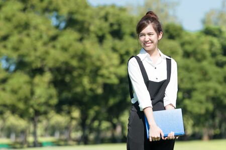 Asian student holding books and smiling while standing in park at collegeの写真素材