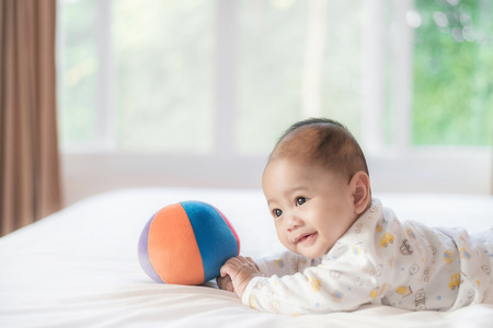 Portrait of 4 month Asian baby lying and playing colorful ball on white bed in bedroom at house.の写真素材
