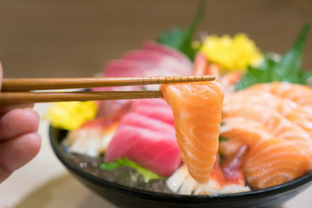 Chopsticks with Salmon sashimi with Mixed sliced fish sashimi on ice in black bowl. Sashimi Salmon Tuna Hamachi Prawn and Surf Calm set, raw fish, japanese food in Asian restuarant.の写真素材