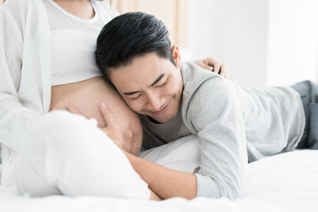 Asian Handsome man is listening to his Asian beautiful pregnant wife's tummy and smiling. Family love concept.の写真素材