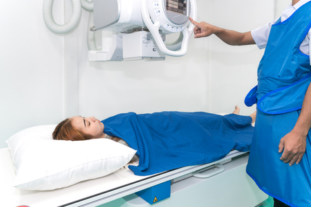 Asian young woman patient lying under X-ray device with radiologist preparing machine in X-ray room at hospital.の写真素材