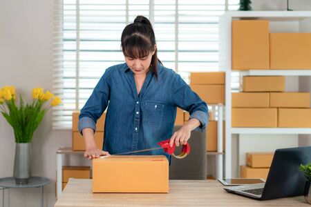 Asian teenager owner business woman work at home for online shopping, wrapping products with brown boxes  for delivery mail shipping with office equipment, entrepreneur lifestyle conceptの写真素材