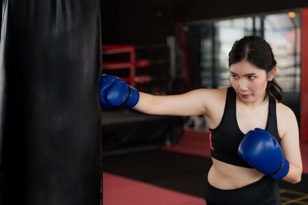 Portrait of Asian confident young boxer woman with blue boxing gloves, punching a Bag in professioal gym. Sporty fit for healthy lifestyle Asian model of boxing gym concept.の写真素材