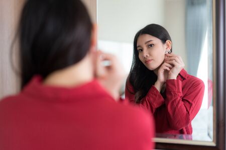 Beautiful Asian Woman wearing red dressed putting star earring looking in mirror in her bedroom at home. Makeup in morning getting ready before going to work.の写真素材