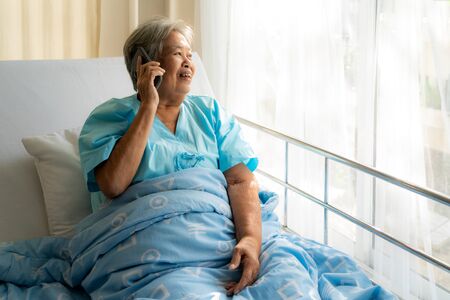 Asian senior elderly using chatting to family on her mobile phone and smiling, with delight as she listens to the conversation while she illness in hospital.の写真素材