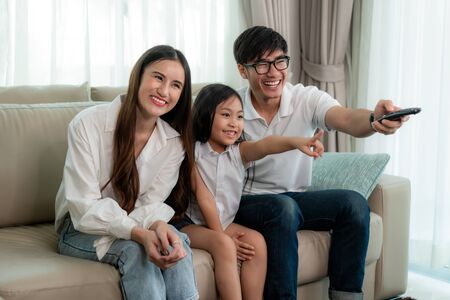 Candid of happy Asian family enjoy weekend activity by watching TV program at home. Father, mother and daughter sitting on sofa table holding remote on hand and pointing toward to TVの写真素材