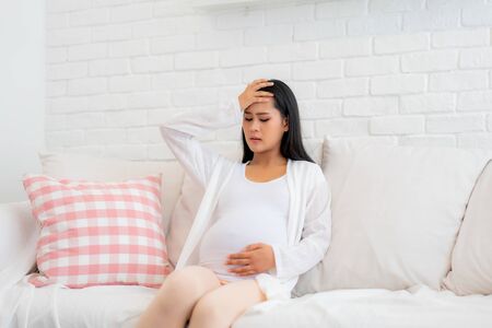 Young pregnant woman with headache sitting on sofa in living room at house. Pregnancy symptoms, expectation, parenthood concept, copy space.
の写真素材
