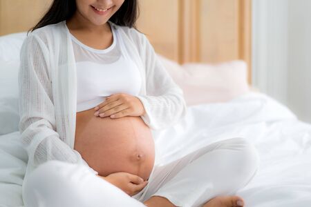 Close up of happy smiling pregnant Asian woman sitting on bed and touching her belly at home. Pregnancy, rest, people and expectation concept の写真素材