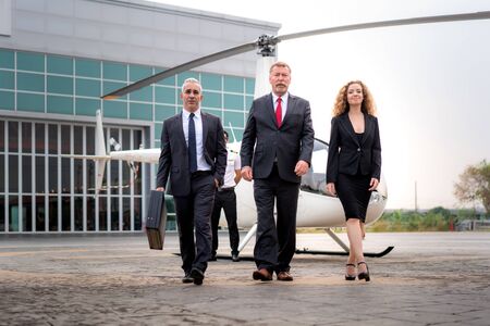 Three Businessman and businesswoman manager with Briefcase walking from helicopter when he arrive destination at helipad in airport with helicopter and captain standing near in background. の写真素材