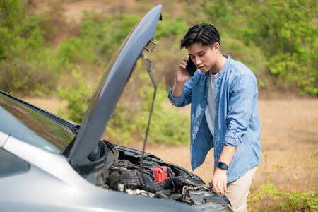 Asian young unhappy man talking on a cell phone  in front of the open hood  broken down car On Country Road Phoning For Help. Broken Car On The Road in During journey.
の写真素材