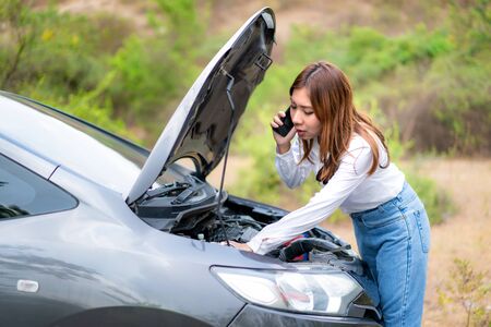 Asian young unhappy woman talking on a cell phone  in front of the open hood  broken down car On Country Road Phoning For Help. Broken Car On The Road in During journey.
の写真素材