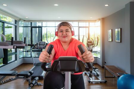 Happy Asian Overweight woman with earphone training on exercise bike in modern gym, happy and smile...