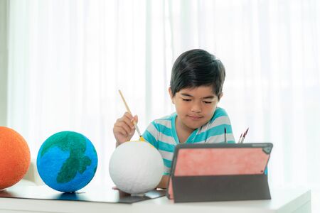 Asian elementary schoolboy painting the moon in science class learning about the solar system via video conference with teacher and other classmates at home, Homeschooling and distance learning.の写真素材
