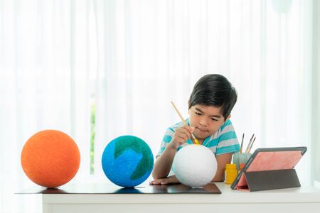 Asian elementary schoolboy painting the moon in science class learning about the solar system via video conference with teacher and other classmates at home, Homeschooling and distance learning.の写真素材
