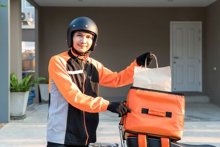 Delivery Asian man wearing orange uniform and ready to send delivering Food bag in front of customer houes with case box on scooter, express food delivery and shopping online concept.の写真素材