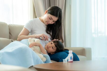 Asian young mother checking measurement temperature of her sick little daughter feel like fever or flu in sofa at home. Visuals of people feeling sick and suffering from common ailments.の写真素材