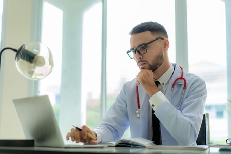 Man doctor is thinking about the result of an examination of his patients. Emotion with the young man doctor being exhausted. Pensive nurse reading some information on the laptop screenの写真素材