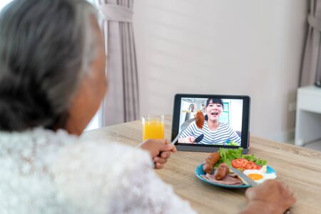 Asian elderly woman virtual happy hour meeting and eating food online together with her daughter in video conference with digital tablet for a online meeting in video call for social distancing.の写真素材