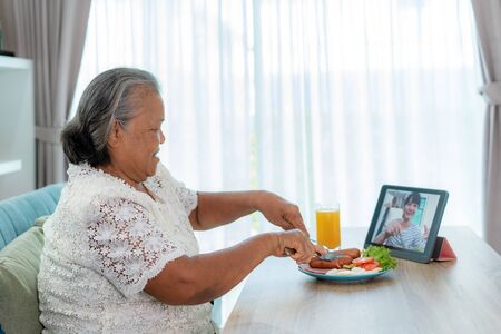 Asian elderly woman virtual happy hour meeting and eating food online together with her daughter in video conference with digital tablet for a online meeting in video call for social distancing.の写真素材