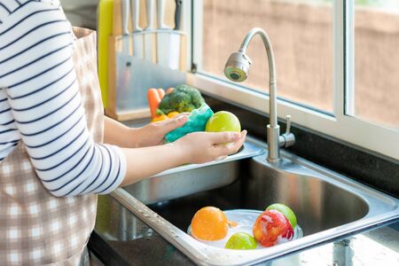 Asian healthy woman washing an apple and other fruit above kitchen sink and applying a fruit / vegetable with soap to eliminate the chances of contamination COVID-19.の写真素材