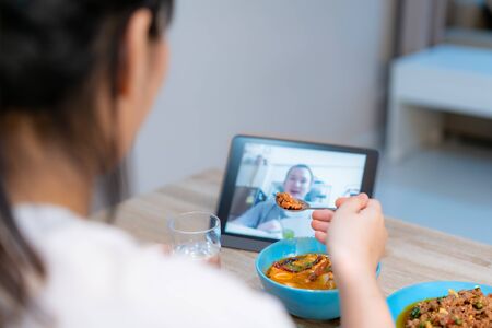 Asian woman virtual happy hour meeting dinner and eating food online together with her boyfriend in video conference with digital tablet for a online meeting in video call for social distancingの写真素材