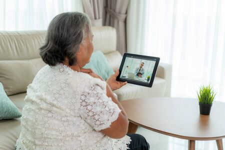 Back view of elderly woman making video call with her doctor with her feeling sore throat on digital tablet online healthcare digital technology service consultation while staying at home.の写真素材