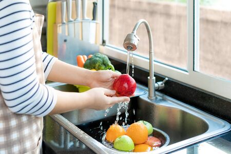 Asian healthy woman washing an apple and other fruit above kitchen sink and cleaning a fruit / vegetable with water to eliminate the chances of contamination COVID-19.の写真素材