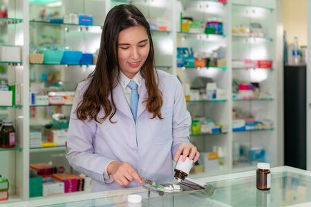 Asian young woman pharmacist pour and count drug capsules on stainless tray, she prepare drug pack to patient in the pharmacy drugstore. Medicine, pharmaceutics, health care and people concept.の写真素材