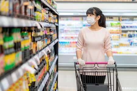 Asian woman with hygienic mask and rubber glove with shopping cart in grocery and looking for sauce or flavoring to buy during covid-19 outbreak for preparation for a pandemic quarantineの写真素材