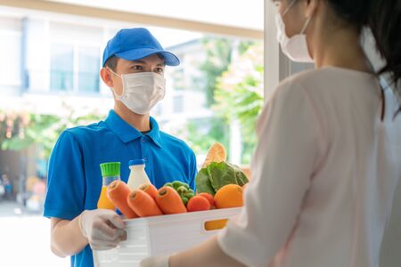 Asian delivery man wearing face mask and glove with groceries box of food, fruit, vegetable and drink give to woman costumer in front of the house during time of home isolation.の写真素材