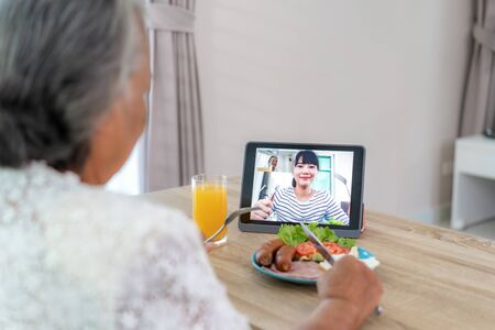 Asian elderly woman virtual happy hour meeting and eating food online together with her daughter in video conference with digital tablet for a online meeting in video call for social distancing.の写真素材