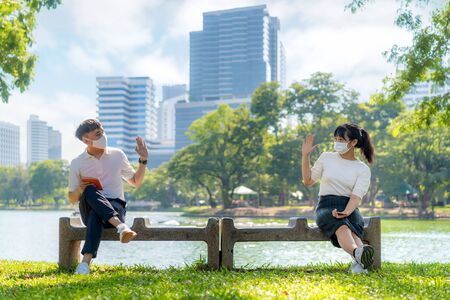 Asian young man and woman greet and say hello with they friend and wearing mask sitting distance of...