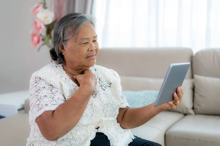 Asian Elderly woman making video call with her doctor with her feeling sore throat on digital tablet online healthcare digital technology service consultation while staying at home.の写真素材