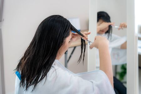 Asian woman cutting own her hair with haircutting scissors at home they stay at home and shelter In place during time of home isolation against Novel coronavirus or COVID-19の写真素材