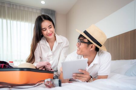 Happy couple packing suitcase on bed in bedroom and lying and looking digital tablet for search and planning travel trip online. Asian backpacker travel lifestyle concept.の写真素材