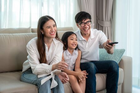 Candid of happy Asian family with father, mother and daughter enjoy weekend activity spending more time at home and therefore probably more time in front of the televisionの写真素材