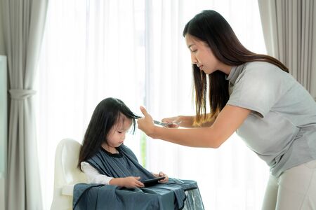 Asian Mother cutting hair to her daughter in living room at home while stay at home safe from Covid-19 Coronavirus during lockdown. Self-quarantine and social distancing concept.の写真素材