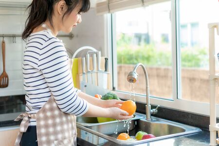 Asian healthy woman washing an orange and other fruit above kitchen sink and cleaning a fruit / vegetable with water to eliminate the chances of contamination COVID-19.の写真素材